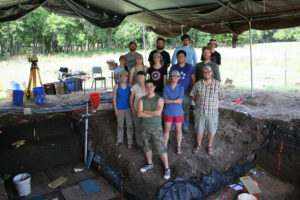 group of male and female students standing under tent at excavation site