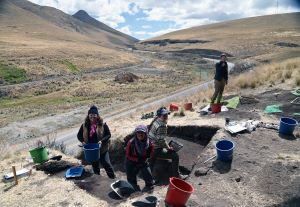 Four archaeologists working on excavation site hillside in the Peruvian highlands. Rolling hills in background with a road running through the middle. Three people are standing in a square excavation hole and one is standing near some buckets on the side. 