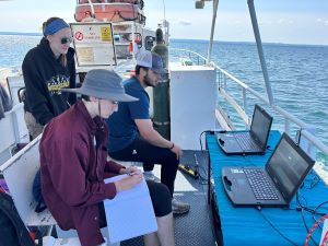 Three Archeological students look at two computers on deck of a large white boat floating on Lake Erie. One is wearing a maroon shirt, one in a blue shirt and one in a black sweatshirt. There are orange lifeboats in the back ground.