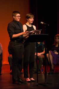 Two performers standing behind podium on stage at The Coming Out Monologues