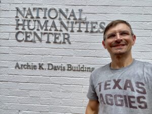 A man in a grey shirt that reads "Texas Aggies" standing in front of a white brick building that reads "National Humanities Center Archie K. Davis Building"