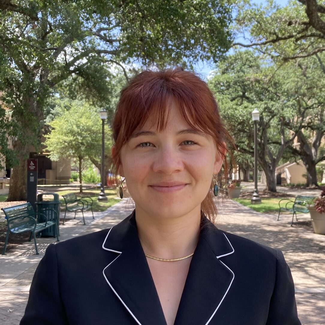 Haley Burke smiling at the camera and wearing a black blazer. Trees and benches can be seen in the background.