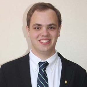 Noah Crawford smiling at the camera and wearing a blue tie, white shirt, and black blazer.