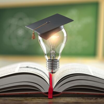 An open book with a red ribbon marking the pages. Above it, a lit lightbulb with a graduation cap placed on top. In the background, blurred chalkboards with unreadable text and a drawing of an atom.
