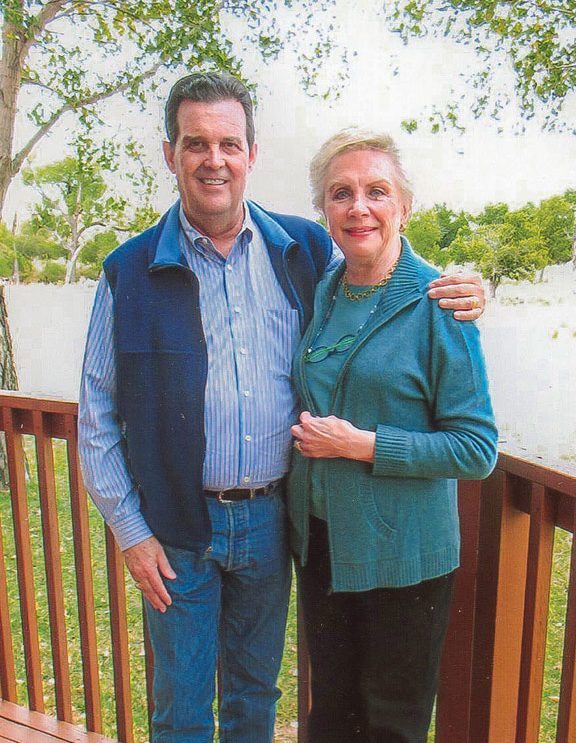 Reverand William Nix '63 stands with his arm around wife Newlyn Nix outside in front of a wooden deck railing surrounded by trees.