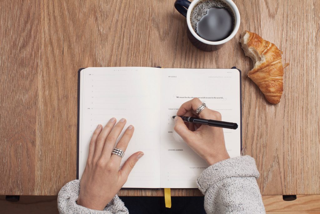 Person writing on a planner next to coffee and croissant.