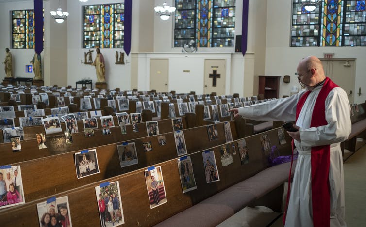 Priest looking at empty pews holding photos of people.