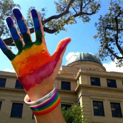 An open hand painted with rainbow stripes is raised in the air with the Academic Building in the background.