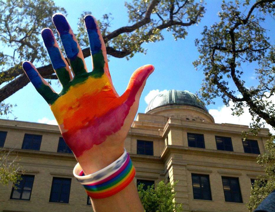An open hand painted with rainbow stripes is raised in the air with the Academic Building in the background.