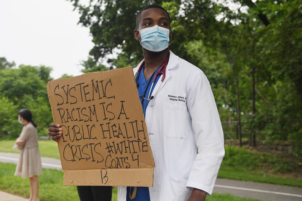 Doctor holding protest sign that says "Systemic racism is a public health crisis #white coats for B"