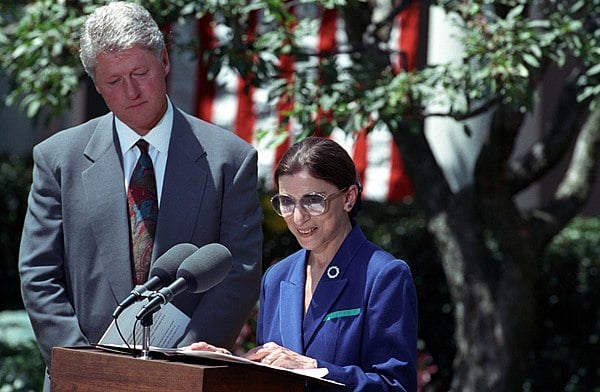 Photo Ruth Bader Ginsburg giving a speech as Bill Clinton looks over her shoulder.