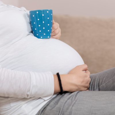 Detail of a pregnant woman's body. Pregnant woman sitting on a couch holding her belly and a cup of tea