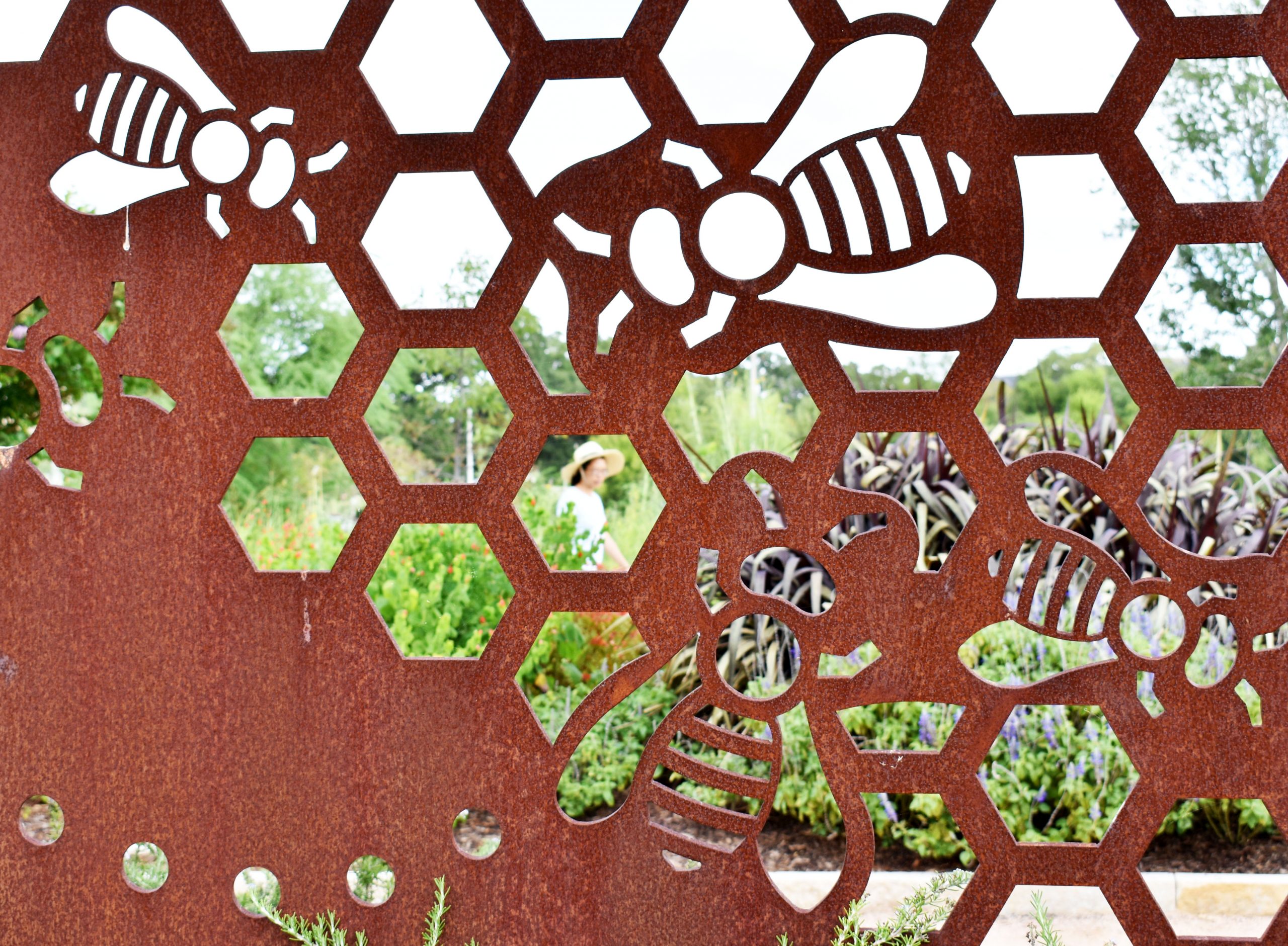 Through metal artwork laser cut to look like an active bee hive, you see a lady in a broad brimmed hat taking a leisurely stroll through The Gardens.