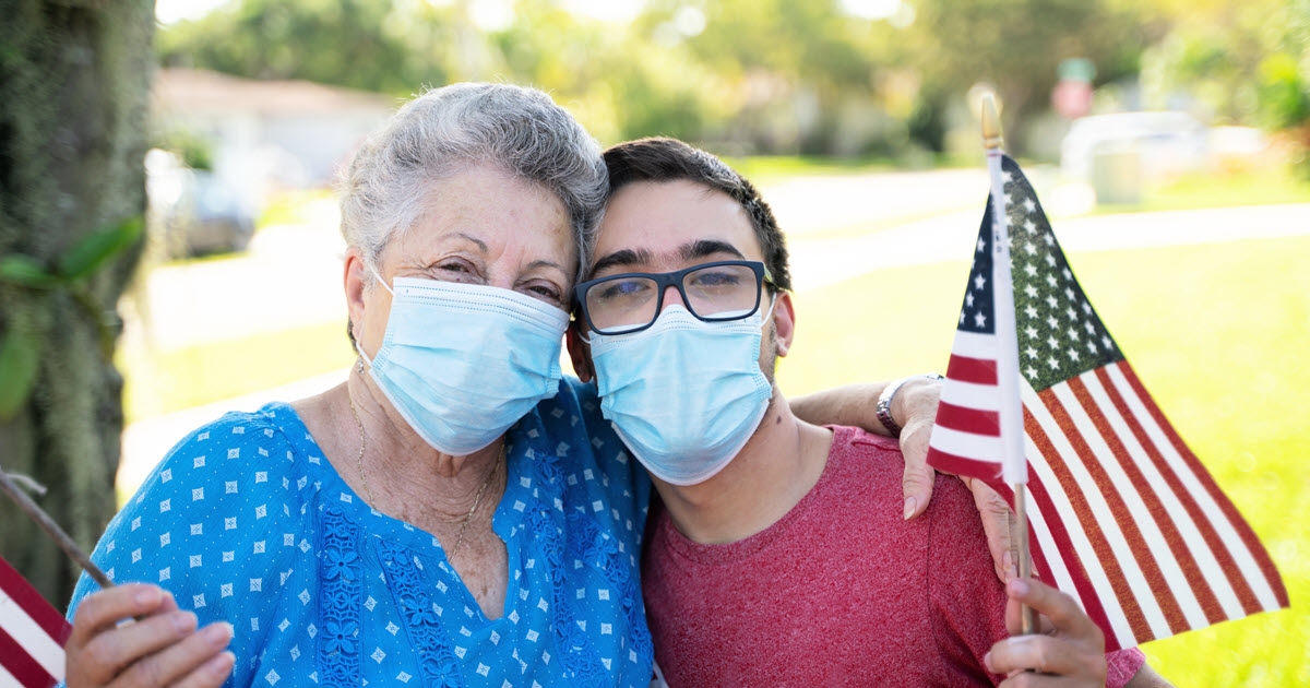 A grandmother and grandson smile through COVID protective masks while sharing a hug and waiving American flags.