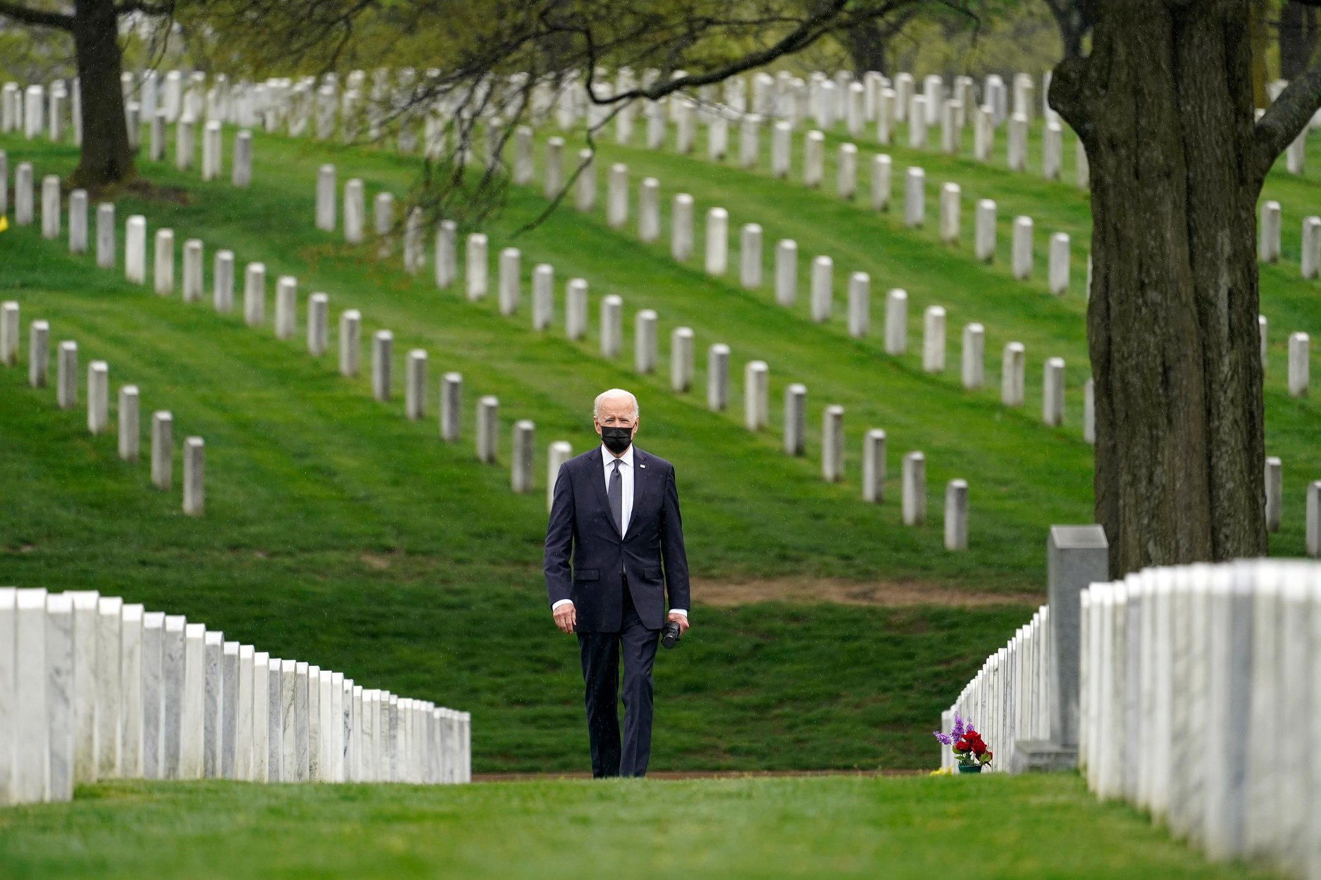 President Joe Biden walks through Arlington National Cemetery. The grass is green, the precision with which the tomb stones are placed is unmistakable. 