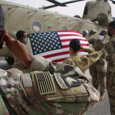 US Army troops salute the United States Flag with an aircraft in the background.