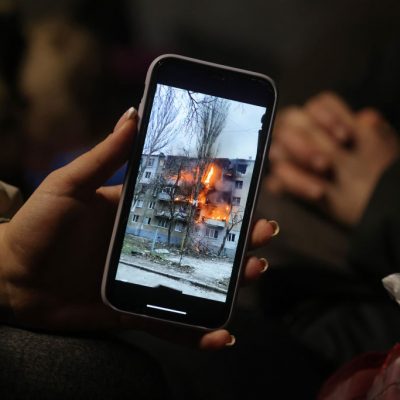 Hand holds phone displaying burning building in Ukraine.